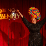 Performer on stage in a black sequined dress and colourful wig with arms raised in front of a Late Night Assembly sign, promoting live comedy and cabaret entertainment.