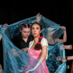 Youth theatre students perform a staged scene, with young actors forming an ensemble around a lead performer, showing drama education, teamwork, and confidence building through performance.