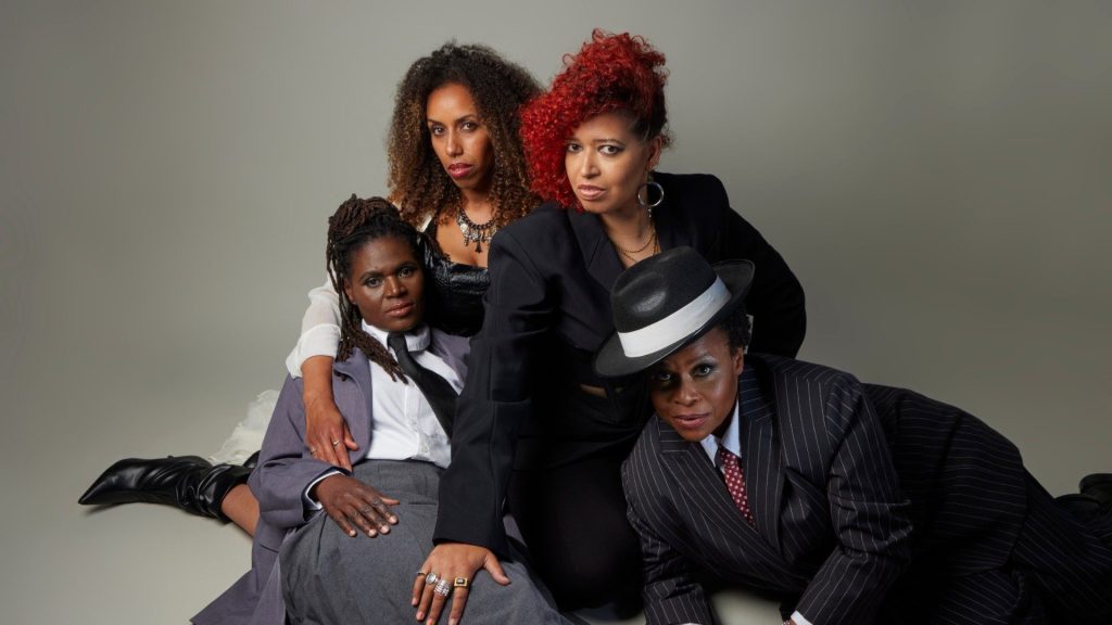 Four black women pose against a neutral background. Two sit in suits, one with a hat, exuding confidence. Two sit behind, dressed stylishly with bold expressions. Mood: Empowering.