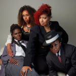 Four black women pose against a neutral background. Two sit in suits, one with a hat, exuding confidence. Two sit behind, dressed stylishly with bold expressions. Mood: Empowering.