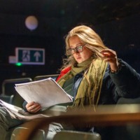 Young woman wearing glasses reads printed notes while seated in a theatre space, preparing materials to lead a youth theatre group and rehearsal activities.