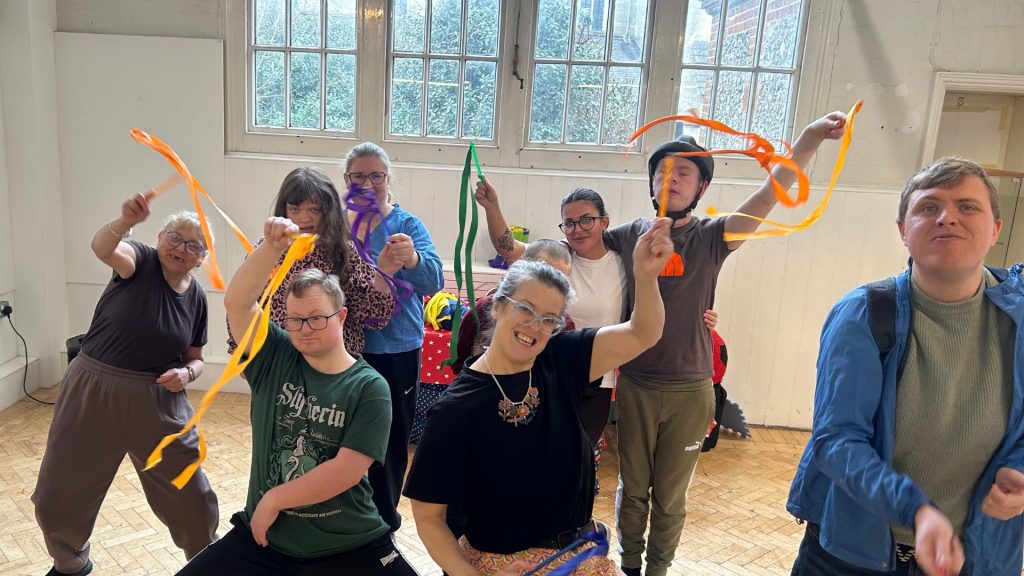 Group of young adults with additional needs and course leader all dancing, smiling and moving with joy. Representing well being for people with learning disabilities and their carers