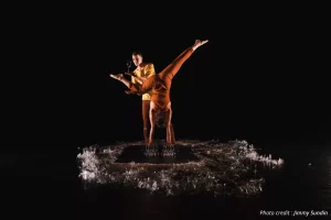 Contemporary circus performers balancing a wine glass during an acrobatic handstand act on a dark stage, showcasing balance, strength and live performance art