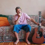 Promotional photo of folk musician Beans on Toast sitting with an acoustic guitar for an upcoming tour.