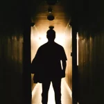 Silhouette of a person walking down a dark hallway toward bright light, illustrating uncertainty, transition, or finding a way forward.